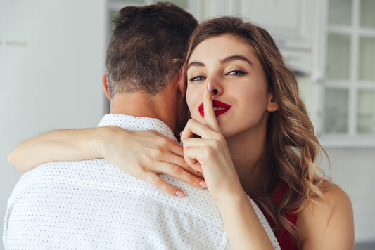 A woman with wavy brown hair and vibrant red lipstick shares an intimate moment with a man in a white dotted shirt. As they embrace, she playfully places her index finger on her lips, signaling a secret or shared mystery.